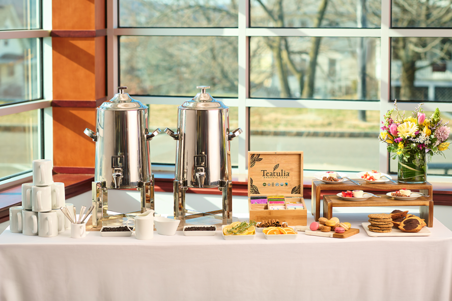 A table with an assortment of beverage and desserts options like a coffee dispenser, tea bags, cups, macarons