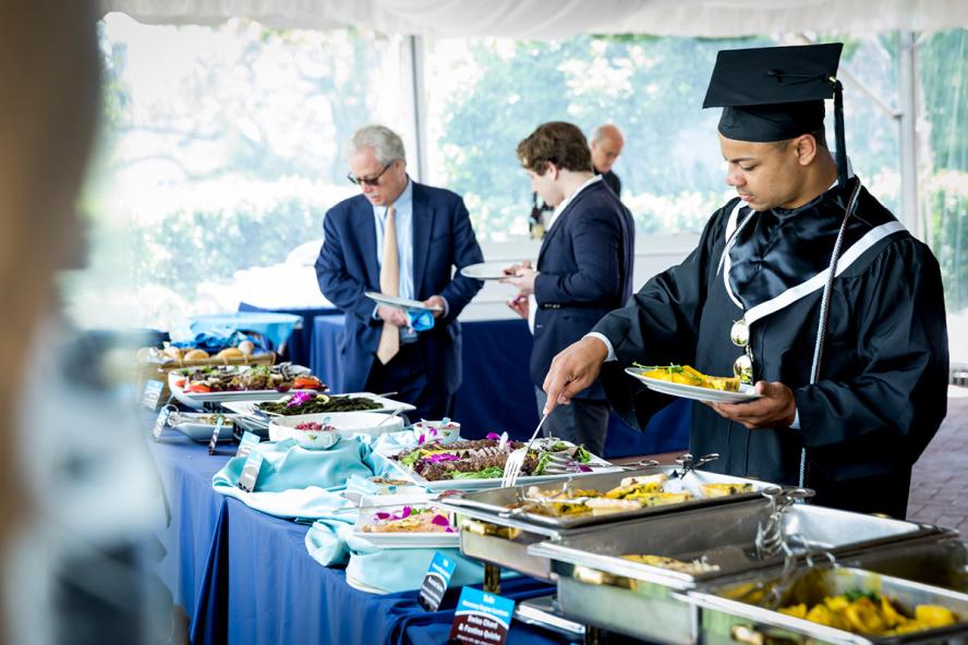 A graduate in cap and gown selects food at a buffet under a tent outdoors